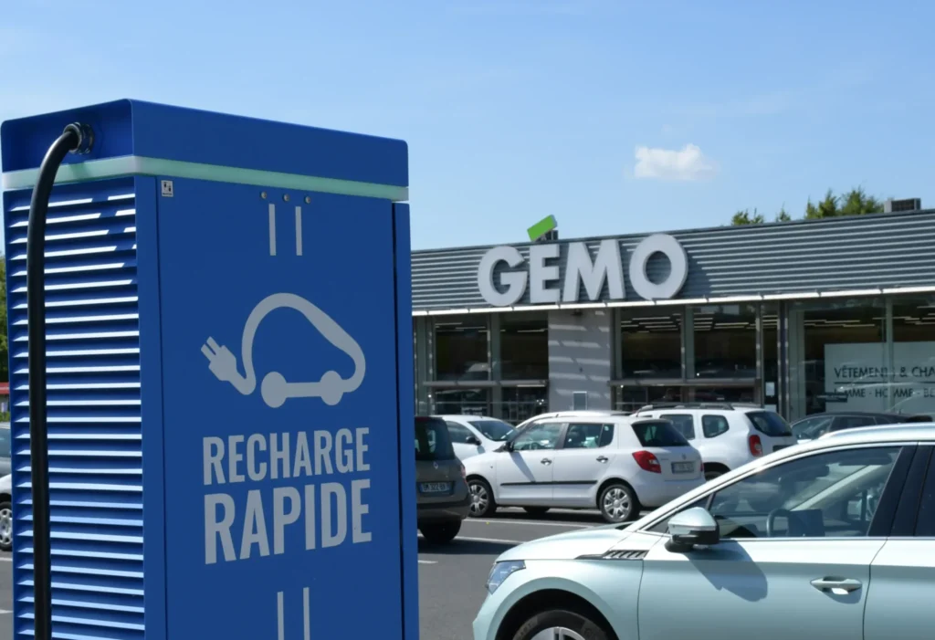 Borne de recharge rapide pour véhicule électrique installée sur le parking d’un centre commercial, voiture électrique stationnée en charge devant un magasin, infrastructure de mobilité durable facilitant la recharge lors des achats