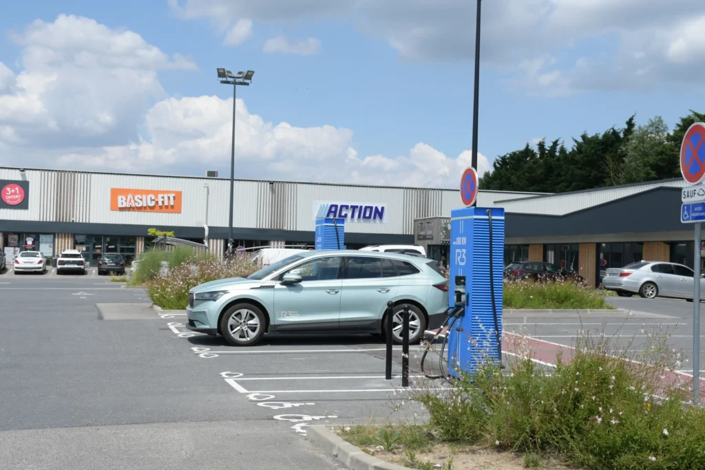 Voiture électrique en charge sur borne rapide dans un parking près des magasins Basic-Fit et Action.